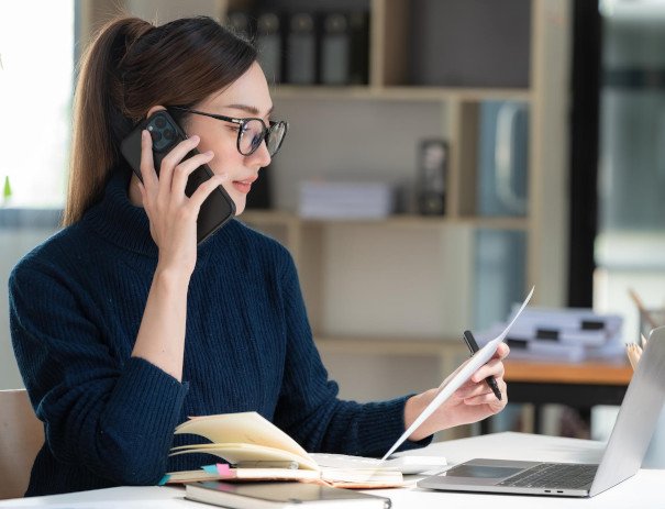 Young woman in a professional setting, multitasking by talking on the phone while reviewing documents. She is seated at a desk with an open notebook, papers, and a laptop, focusing on her work in a modern office environment.